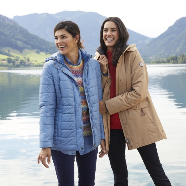 Zwei Frauen stehen an einem See in den Bergen und tragen Jacken.