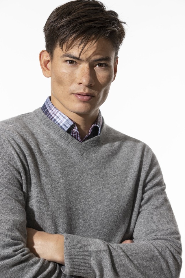 Ein Mann mit einem grauen Cashmere Pullover und einem Hemd darunter schaut in die Kamera