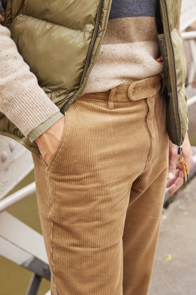 Person in beiger Cordhose, Strickpullover und grüner Weste steht draußen mit Händen in den Taschen.