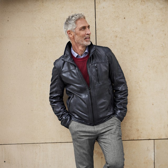 Mann mit einer schwarzen Lederjacke steht an einer Wand und lacht.