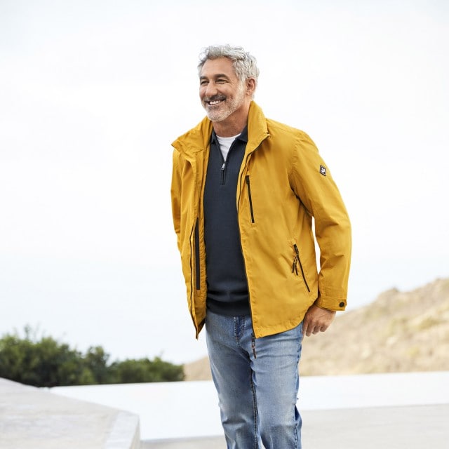 Ein Mann mit gelber Jacke und Jeans steht draußen und lacht