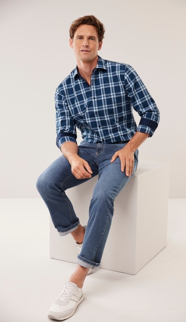 Mann in kariertem Hemd und Jeans sitzt auf weißem Würfel, trägt weiße Sneaker.