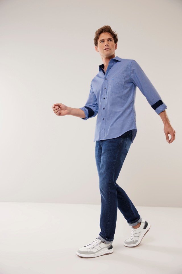 Mann in blauem Hemd und Jeans geht in einem hellen Raum, trägt weiße Sneaker.