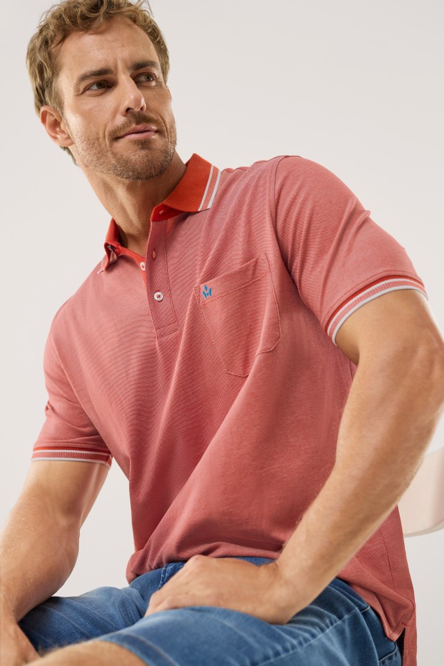 Ein Mann trägt ein rotes, kurzärmliges Poloshirt mit Brusttasche und einem orange abgesetzten Kragen sowie blauer Jeans-Shorts.