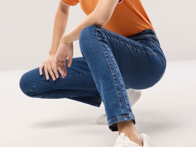 Eine Frau hockt am Boden und trägt eine blaue Jeans, ein orangefarbenes Oberteil und weiße Sneaker.