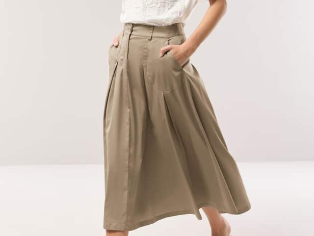 Eine Frau trägt einen weit schwingenden, knielangen Rock in Beige und hat die Hände in den Taschen.