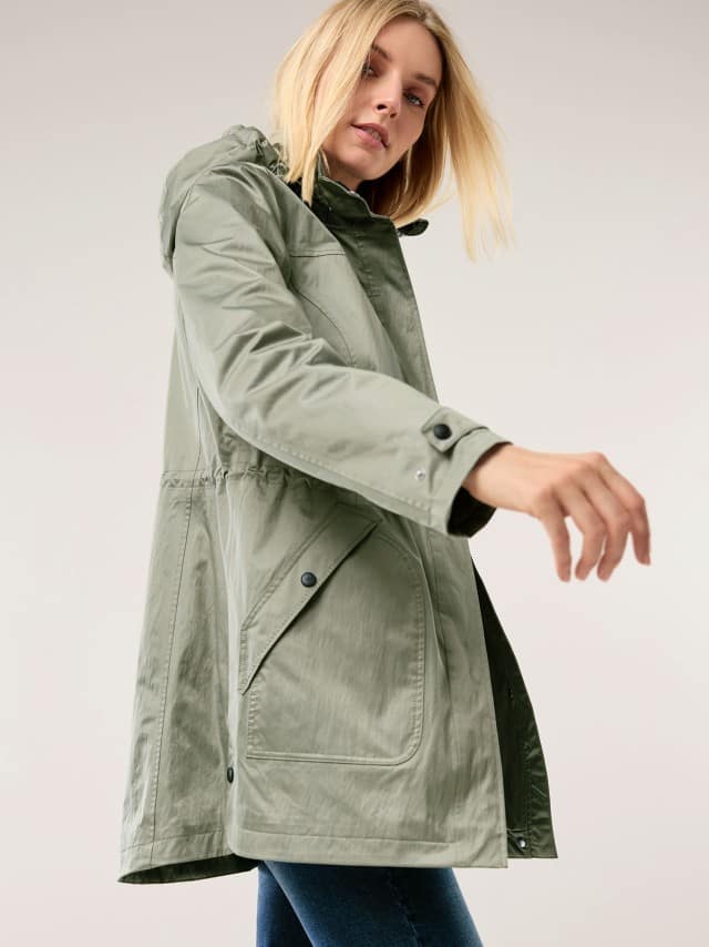 Eine Frau trägt eine hellgrüne, längere Jacke mit Kapuze und großen Taschen und bewegt einen Arm leicht nach vorn. Eine Frau trägt eine hellgrüne, längere Jacke mit Kapuze und großen Taschen und bewegt einen Arm leicht nach vorn.