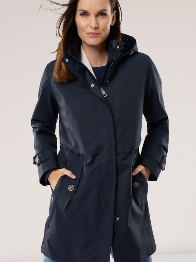 Eine Frau trägt eine dunkelblaue, lange Jacke mit Kapuze und hat die Hände in den Taschen. Eine Frau trägt eine dunkelblaue, lange Jacke mit Kapuze und hat die Hände in den Taschen.