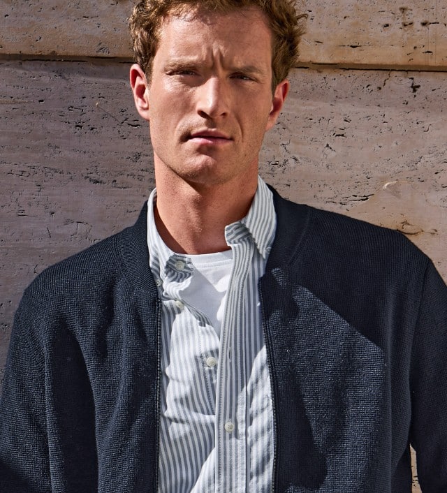 Ein Mann vor einer Natursteinmauer, trägt ein gestreiftes Hemd und darüber einen Cardigan in Marine.