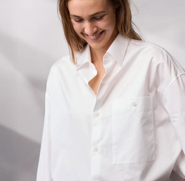 Eine Frau trägt ein weites, weißes Hemd und blickt lächelnd nach unten.