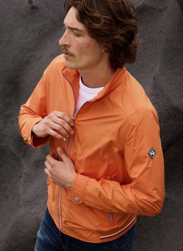 Ein Mann mit mittellangem braunem Haar und Schnurrbart trägt eine orangefarbene Jacke über einem weißen T-Shirt und schließt den Reißverschluss.