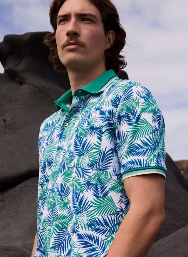 Ein Mann mit mittellangem braunem Haar und Schnurrbart trägt ein weißes Poloshirt mit blau-grünem Palmblattmuster und grünem Kragen vor einer felsigen Küstenkulisse.