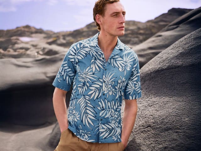 Mann in blauem Hawaiihemd mit weißem Blumenmuster und beigefarbener Hose vor einer felsigen Berglandschaft.