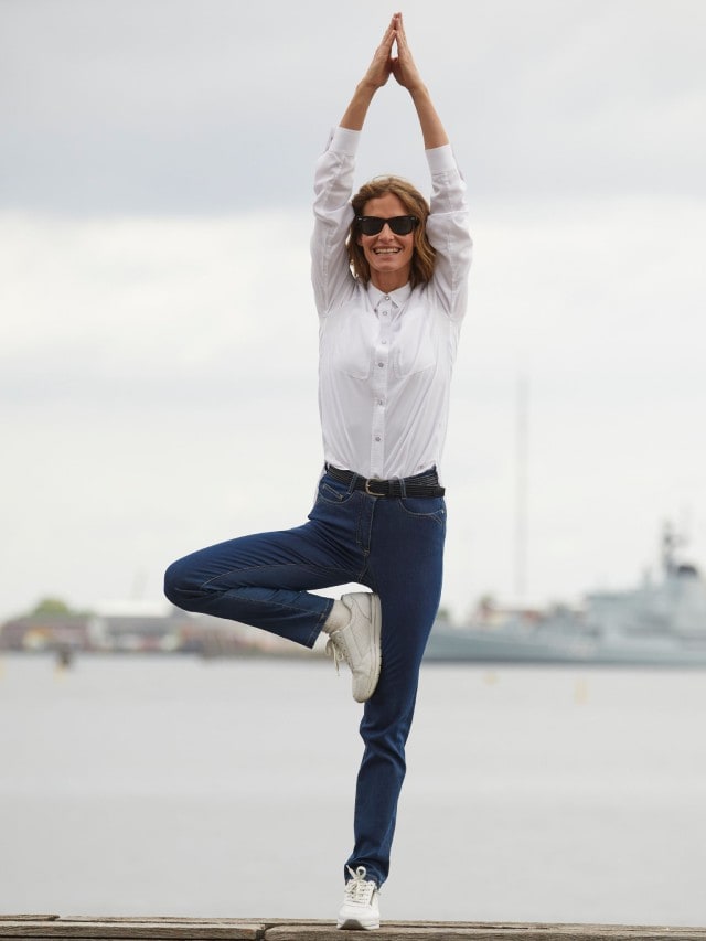 Frau macht eine Yoga Pose in einer blauen Jeans und weißen Bluse am Wasser.