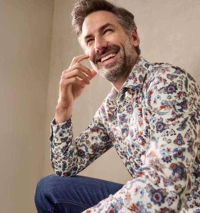 Ein Mann trägt ein helles Hemd mit buntem Blumenmuster und eine Jeans. Der Stil wirkt modisch, lebendig und zugleich entspannt.