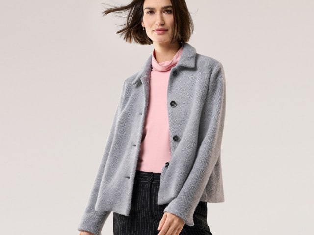 Eine Frau trägt einen grauen, weichen Blazer mit Knöpfen über einem rosa Rollkragen-Shirt. Dazu kombiniert sie eine dunkle Hose mit feinen Streifen, was einen modernen und femininen Look ergibt.