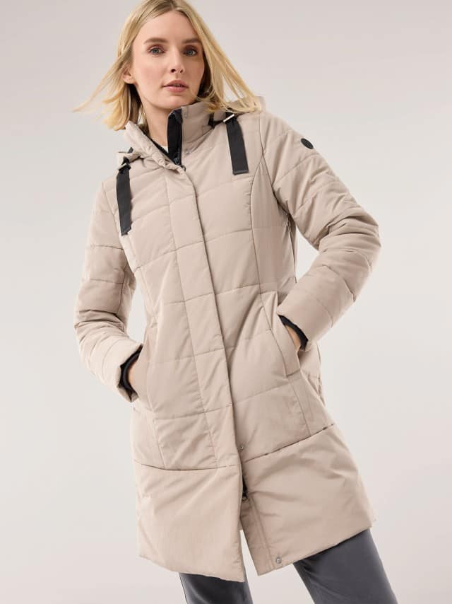 Eine Frau trägt eine hellbeige, gesteppte Winterjacke mit Kapuze und schwarzen Riemendetails. Der Stil wirkt sportlich und leger zugleich. Eine Frau trägt eine hellbeige, gesteppte Winterjacke mit Kapuze und schwarzen Riemendetails. Der Stil wirkt sportlich und leger zugleich.