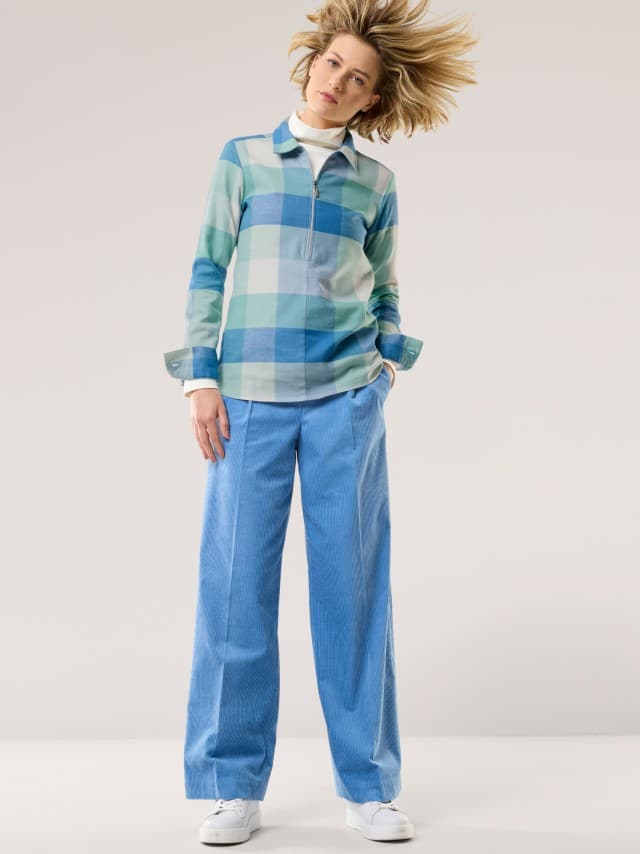 Eine Frau trägt ein kariertes Oberteil in Blau- und Grüntönen über einem weißen Rollkragen. Dazu kombiniert sie weite, hellblaue Cordhosen und weiße Sneaker für einen modernen, frischen Look. Eine Frau trägt ein kariertes Oberteil in Blau- und Grüntönen über einem weißen Rollkragen. Dazu kombiniert sie weite, hellblaue Cordhosen und weiße Sneaker für einen modernen, frischen Look.