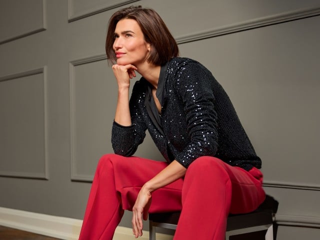 Eine Frau sitzt auf einem Stuhl, ist nach vorne gelehnt und trägt eine schwarze Paillettenjacke, dazu eine rote Hose.