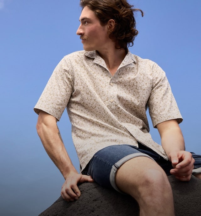 Zwei Männer unter blauem Himmel: Einer trägt ein weißes T-Shirt, beige Shorts und Sonnenbrille, der andere ein gemustertes Hemd und Jeansshorts auf einem Felsen.