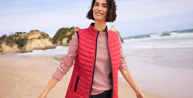 Frau in einem rot-weiß gestreiften Halbarm-Oberteil, roter Steppweste und Jeans läuft lächelnd am Strand entlang und schaut in die Kamera.