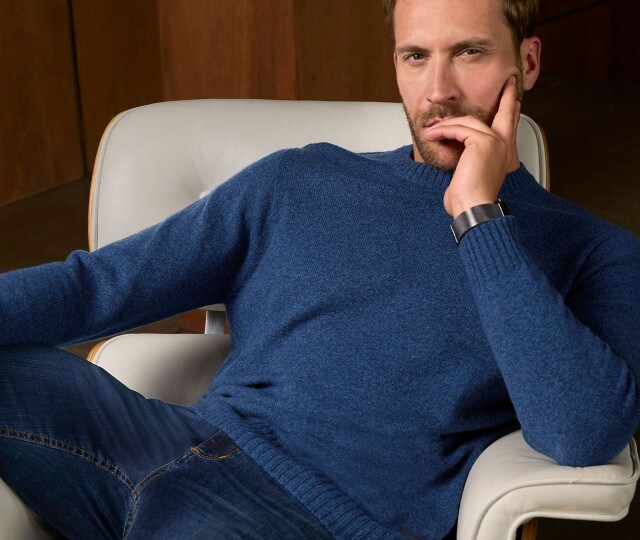 Herr sitzt auf einem Sessel und trägt einen blauen Wollpullover.