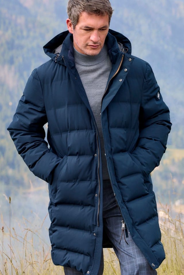 Ein Mann trägt eine lange, dunkelblaue Steppjacke über einem grauen Rollkragenpullover. Das Outfit wirkt funktional, elegant und ideal für kühle Tage.