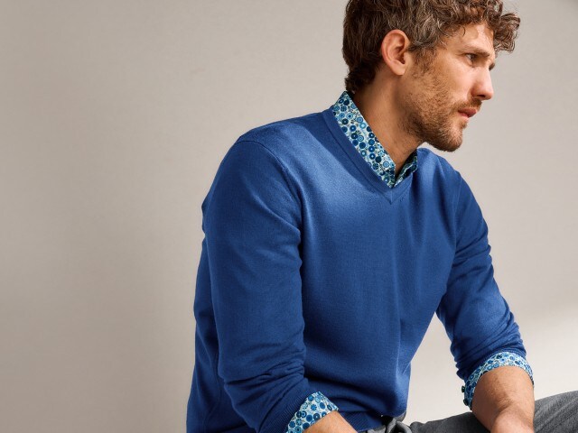 Ein Mann trägt einen dunkelblauen Pullover mit Rundhalsausschnitt, darunter ein gemustertes Hemd.