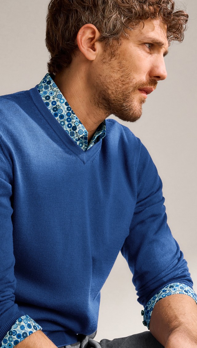 Ein Mann in einem blau gemusterten Hemd unter einem blauen Pullover sitzt vor einem neutralen Hintergrund und schaut zur Seite.