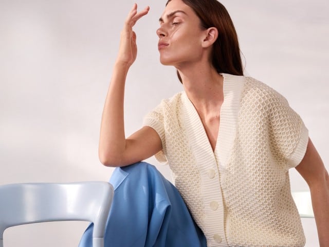 Eine Person sitzt auf einem Stuhl und trägt eine cremefarbene Strickjacke sowie eine hellblaue Hose, während sie mit geschlossenen Augen eine Hand vor das Gesicht hält. Eine Person sitzt auf einem Stuhl und trägt eine cremefarbene Strickjacke sowie eine hellblaue Hose, während sie mit geschlossenen Augen eine Hand vor das Gesicht hält.