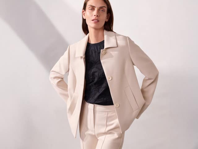 Eine Person steht vor einer neutralen Wand und trägt einen hellen Anzug mit Jacke und Hose, kombiniert mit einem dunklen Oberteil. Eine Person steht vor einer neutralen Wand und trägt einen hellen Anzug mit Jacke und Hose, kombiniert mit einem dunklen Oberteil.