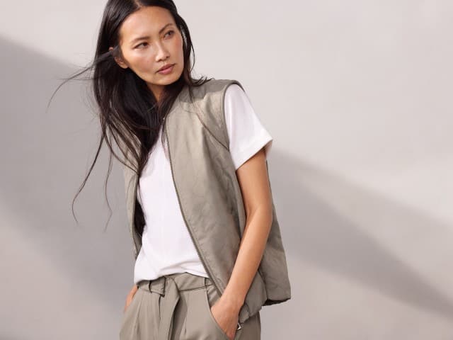 Eine Person trägt ein weißes T‑Shirt, eine leichte gesteppte Weste und eine locker sitzende beigefarbene Hose, während sie zur Seite blickt. Eine Person trägt ein weißes T‑Shirt, eine leichte gesteppte Weste und eine locker sitzende beigefarbene Hose, während sie zur Seite blickt.