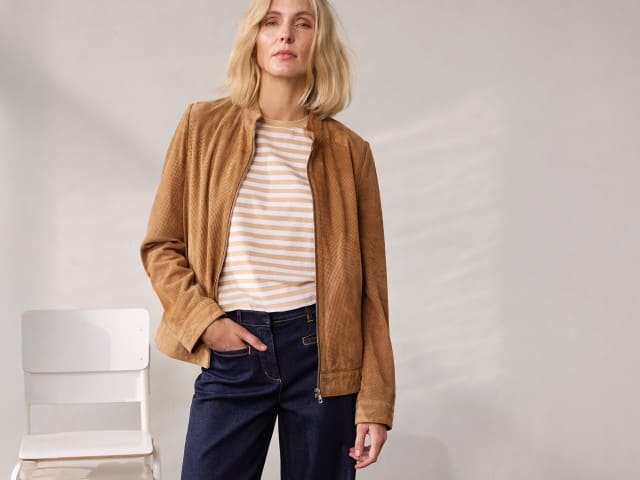 Eine Person steht vor einer neutralen Wand und trägt eine braune Jacke, ein gestreiftes Oberteil und eine dunkelblaue Jeans. Eine Person steht vor einer neutralen Wand und trägt eine braune Jacke, ein gestreiftes Oberteil und eine dunkelblaue Jeans.