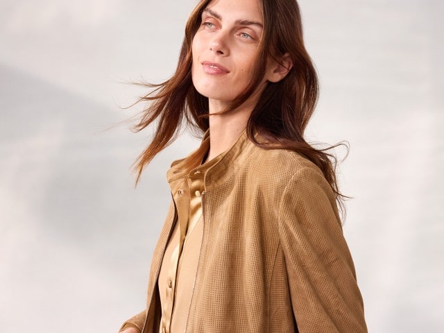 Eine Person trägt eine hellbraune Bluse und eine passende perforierte Jacke und blickt leicht lächelnd zur Seite. Eine Person trägt eine hellbraune Bluse und eine passende perforierte Jacke und blickt leicht lächelnd zur Seite.