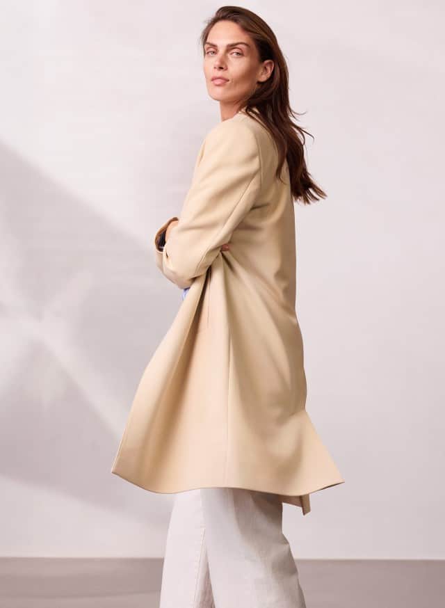 Eine Person trägt einen langen, hellen Mantel über einer weiten, hellen Hose und blickt über die Schulter zur Seite. Eine Person trägt einen langen, hellen Mantel über einer weiten, hellen Hose und blickt über die Schulter zur Seite.