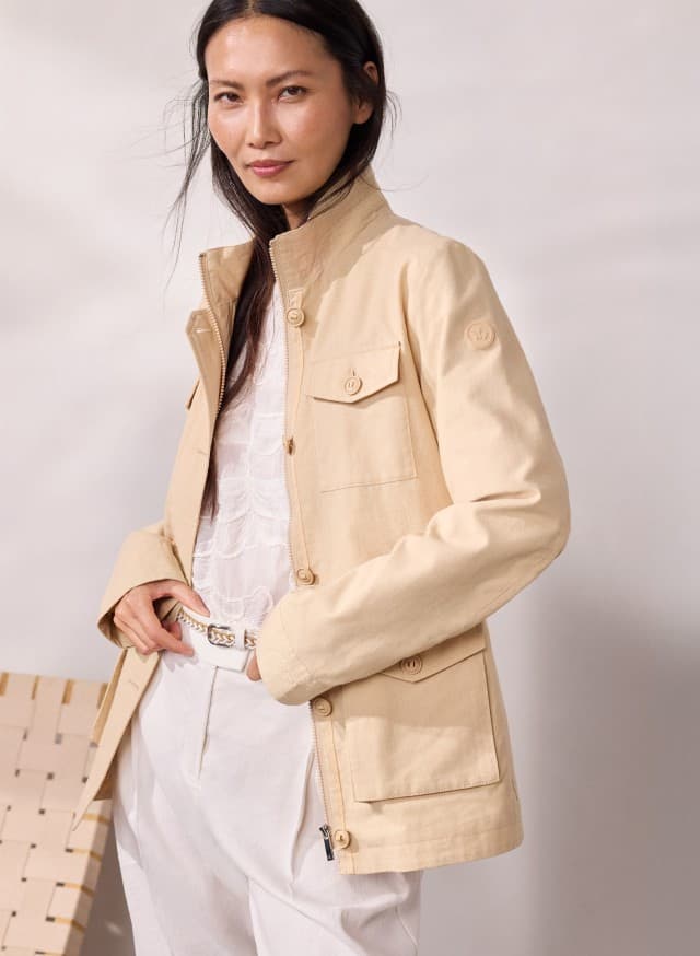 Eine Person trägt eine helle, beigefarbene Jacke über einem weißen, gemusterten Top und einer weißen Hose mit Gürtel. Eine Person trägt eine helle, beigefarbene Jacke über einem weißen, gemusterten Top und einer weißen Hose mit Gürtel.