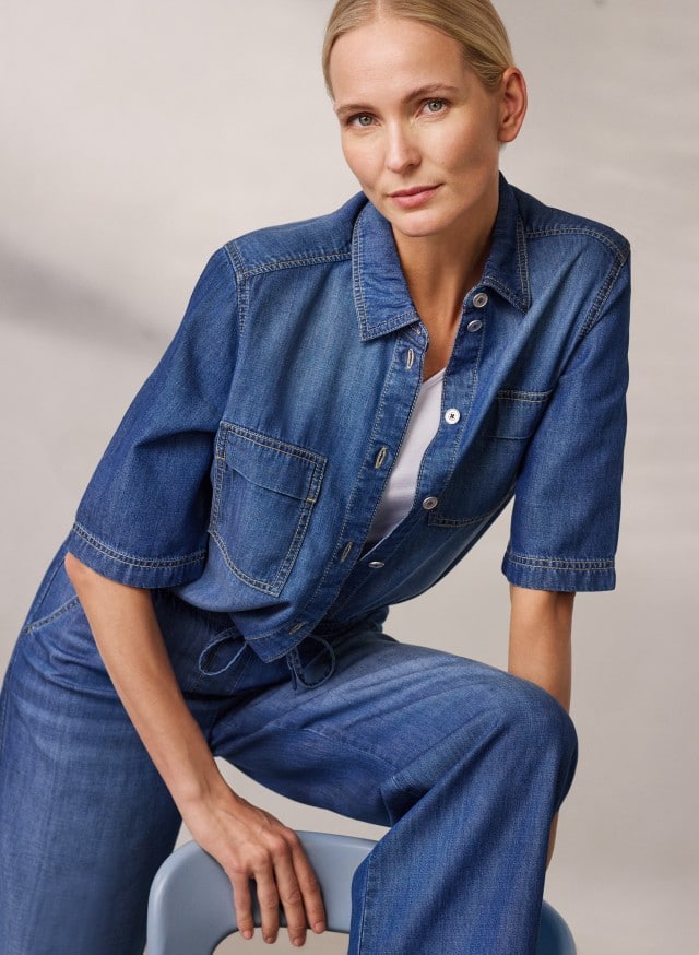 Eine Person trägt ein kurzärmliges Jeanshemd über einem weißen Top sowie eine passende weite Jeanshose und sitzt auf einem Stuhl. Eine Person trägt ein kurzärmliges Jeanshemd über einem weißen Top sowie eine passende weite Jeanshose und sitzt auf einem Stuhl.