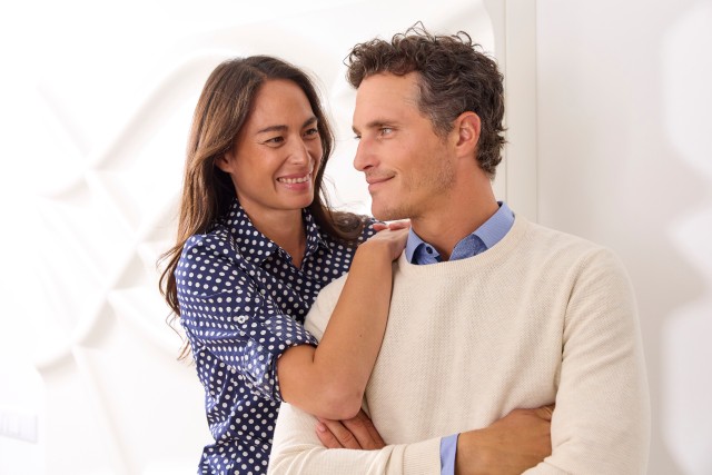 Mann trägt einen hellen Pullover und wird von einer Frau umarmt, die eine Bluse trägt.
