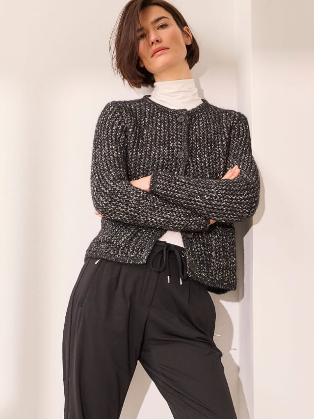 Eine Person trägt einen weißem Rollkragenpullover unter einer grob gestrickten, schwarz-grauen Strickjacke und kombiniert dazu eine schwarze Hose mit Kordelzug.