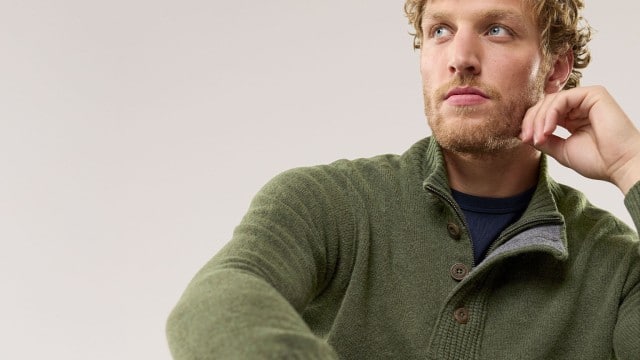 Ein Mann trägt einen olivgrünen Strickpullover mit Knopfleiste und hohem Kragen. Der Stil wirkt gemütlich, natürlich und sportlich-elegant.
