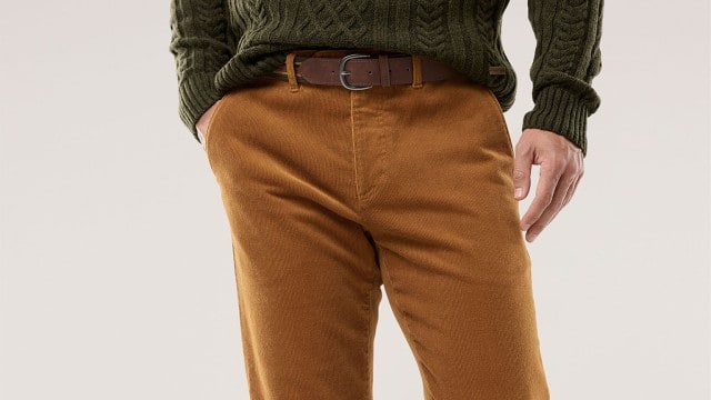 Eine Person trägt eine senffarbene Cordhose mit braunem Gürtel und einen dunkelgrünen Strickpullover. Der Stil wirkt herbstlich, warm und lässig.