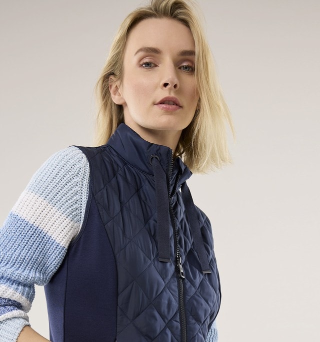 Frau in gestepptem, navyblauen Gilet über gemustertem Pullover und Jeans, vor hellem Hintergrund.