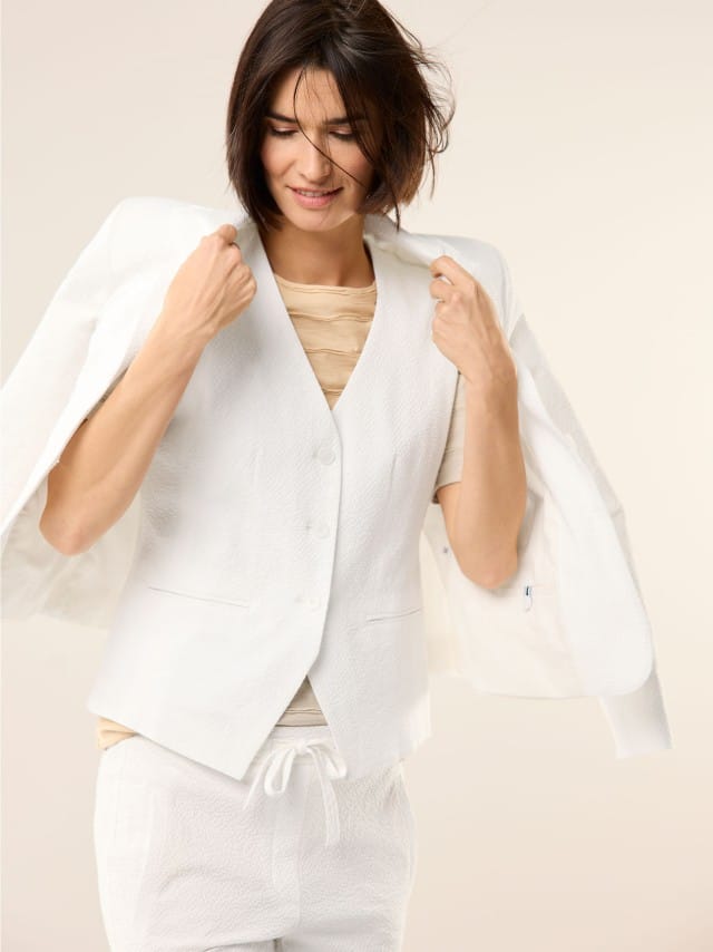 Eine Frau trägt ein locker geschnittenes, weißes Outfit aus Weste, Jacke und Hose über einem beigefarbenen Oberteil, wodurch der Look leicht, modern und entspannt wirkt.