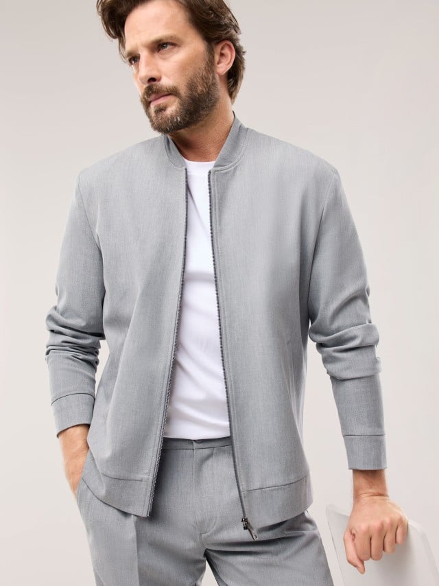Ein Mann trägt ein hellgraues, modern geschnittenes Set aus Jacke und Hose über einem weißen Shirt, was einen klaren, frischen und ruhig‑lässigen Eindruck vermittelt.