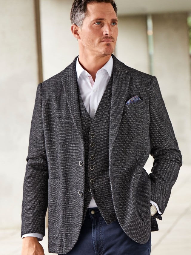 Ein Mann trägt eine dunkel strukturierte Jacke mit passender Weste zu einem weißen Hemd, was gepflegt, modern und souverän wirkt.