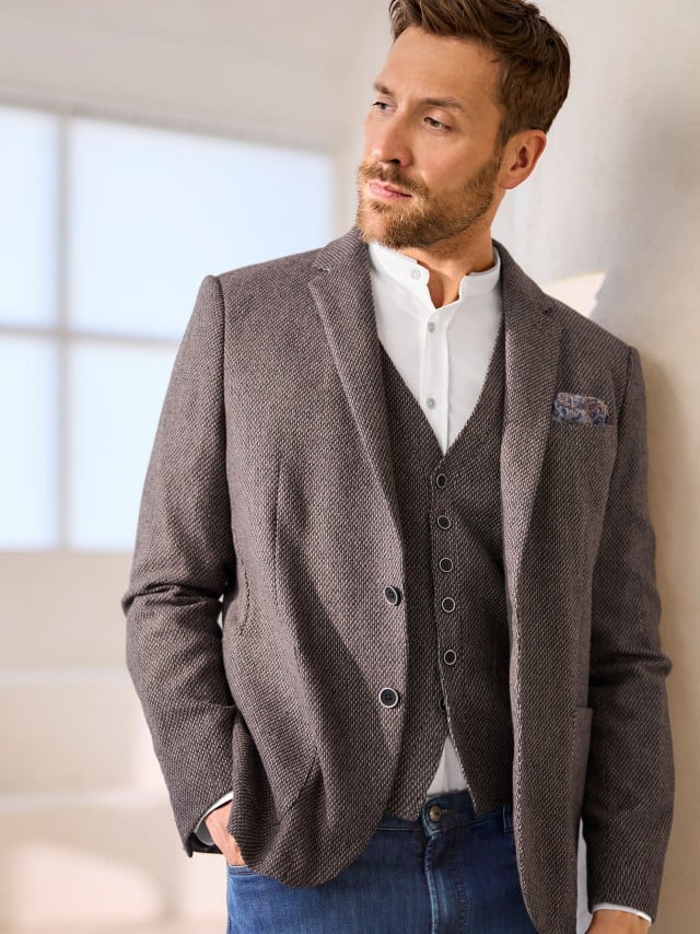Ein Mann trägt eine braun strukturierte Jacke mit passender Weste zu einem weißen Hemd und Jeans, was modern‑lässig und ruhig‑stilvoll wirkt.