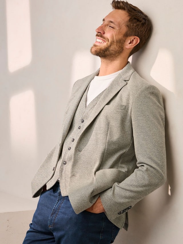 Ein Mann trägt eine hell strukturierte Jacke mit passender Weste zu Jeans und weißem Shirt, was modern‑lässig wirkt und eine warme, entspannte Stimmung ausstrahlt.
