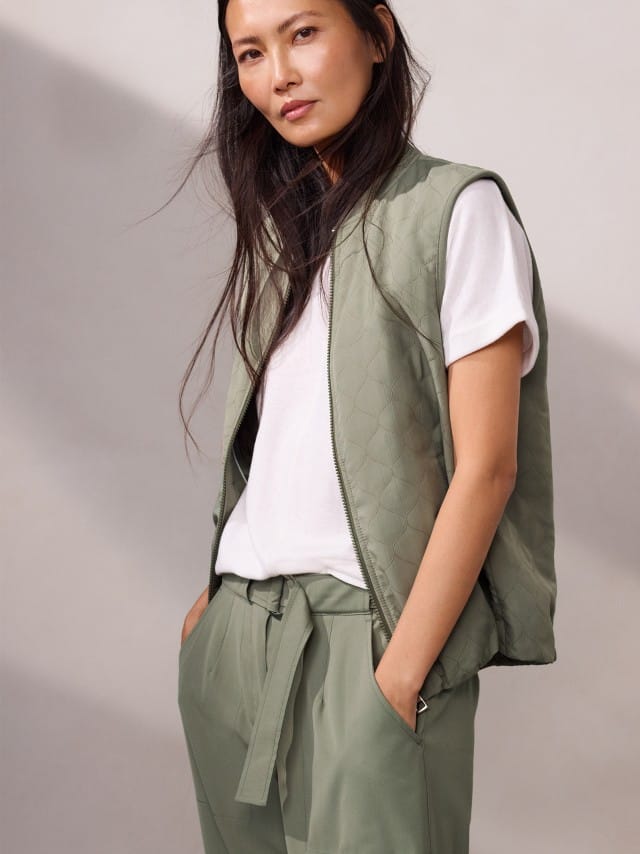 Eine Frau trägt eine leichte, gesteppte Weste zu weißem T‑Shirt und locker gebundenen Stoffhosen, was entspannt, modern und angenehm lässig wirkt.