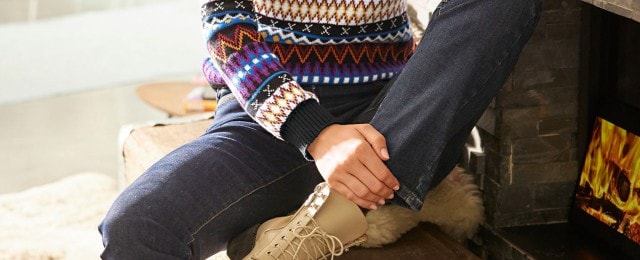 Eine Person sitzt in einem gemütlichen Raum neben einem Kamin. Sie trägt einen bunten Strickpullover mit aufwendigem Muster, dunkle Jeans und helle Schnürstiefel.
