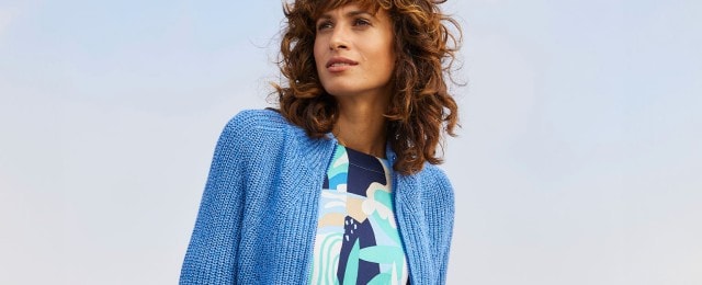 Eine Frau trägt eine blaue Strickjacke über einem gemusterten Oberteil mit Blau-, Grün- und Beigetönen. Der Himmel ist im Hintergrund zu sehen.